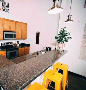 A kitchen with wooden cabinets, a granite countertop, stainless steel appliances, and yellow bar stools. - Cozy mill loft w/ 2 king beds, gym & pool (Spartanburg)