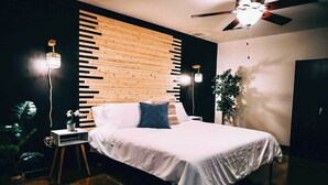 A modern bedroom with a wooden slat wall, a bed with white bedding, two bedside lamps, and a ceiling fan. - Cozy mill loft w/ 2 king beds, gym & pool (Spartanburg)