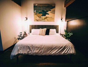 Una cama bien tendida con cabecera, dos lámparas de noche y un cuadro abstracto colgado en la pared.