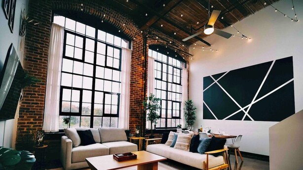 A modern living room with a brick wall, large windows, and a geometric wall art piece. - Cozy mill loft w/ 2 king beds, gym & pool (Spartanburg)