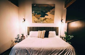 A bedroom with a large bed, two bedside lamps, a wall-mounted artwork, and potted plants.