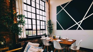A dining area with a brick wall, large windows, and a modern geometric wall art piece. - Cozy mill loft w/ 2 king beds, gym & pool (Spartanburg)