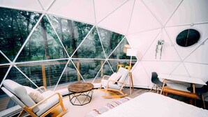 Un interior moderno con diseño geométrico, una ventana de vidrio grande, un sofá blanco, una mecedora de madera, una mesita redonda pequeña y una cama blanca.