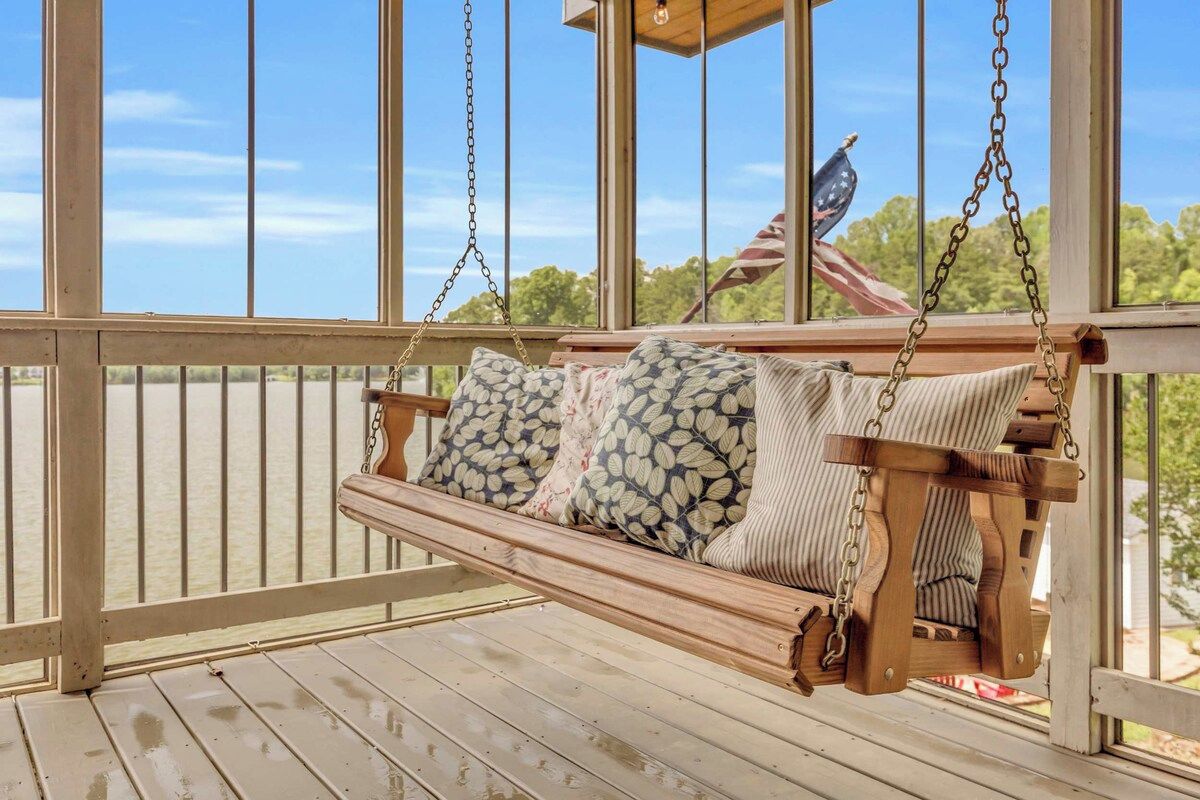 A wooden swing with cushions, a wooden railing, and a view of trees and an American flag.