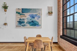 Un coin repas avec une table et des chaises en bois, un grand tableau abstrait accroché au mur et une fenêtre donnant sur l’extérieur.
