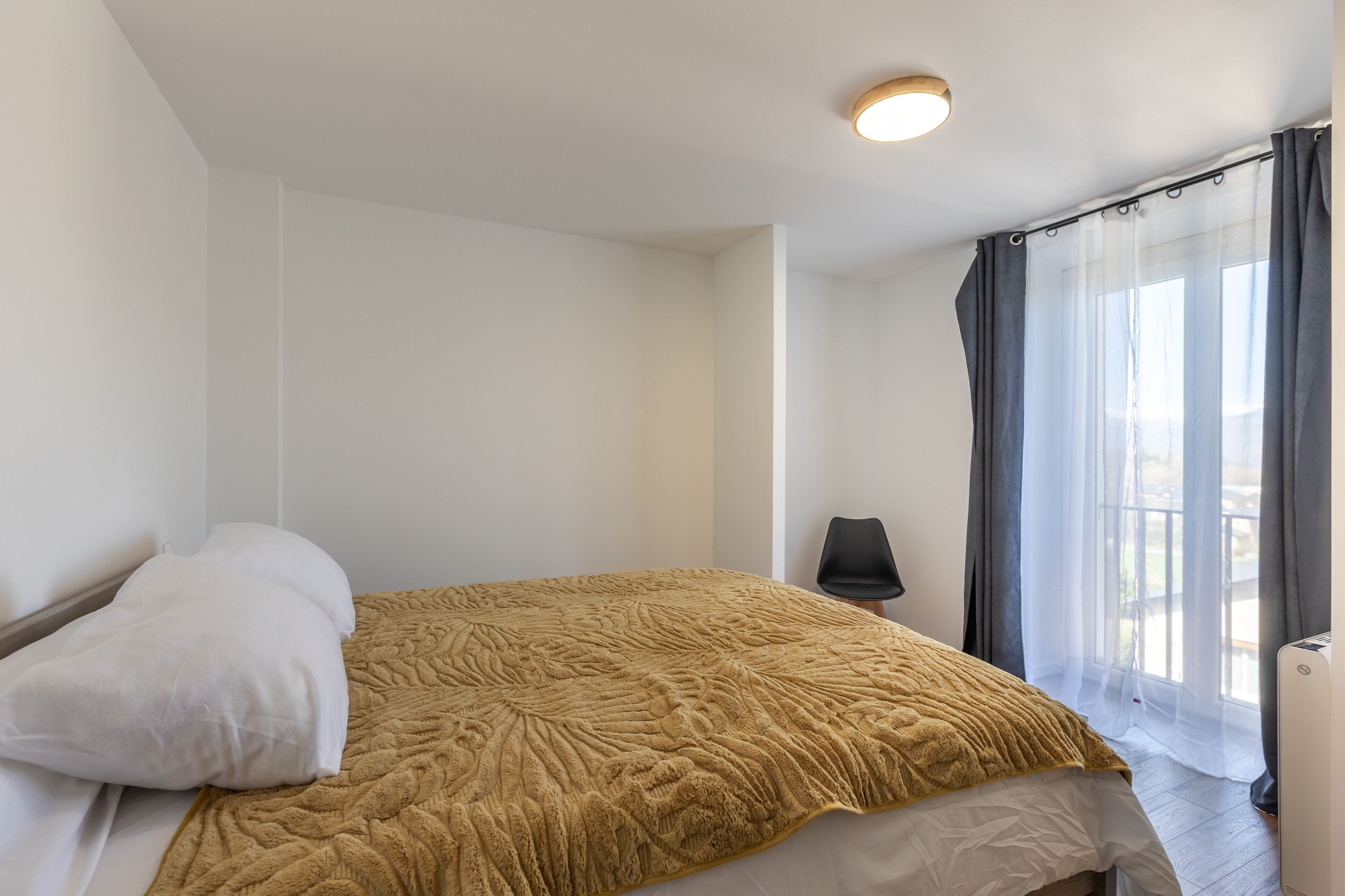 A neatly made bed with a textured blanket, a white pillow, and a black chair beside a window with sheer curtains.