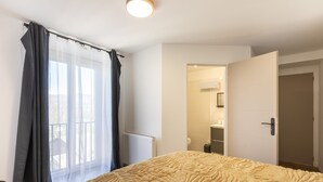Un dormitorio con una cama grande, una ventana con cortinas traslúcidas, una puerta que da a un baño y una lámpara en el techo.