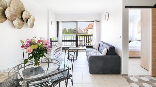 Een moderne woonkamer met een glazen eettafel, een bank en een balkon met uitzicht op huizen.