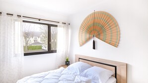 Een slaapkamer met een bed, een raam met gordijnen, een ventilator aan de muur en een tafeltje met een plant.