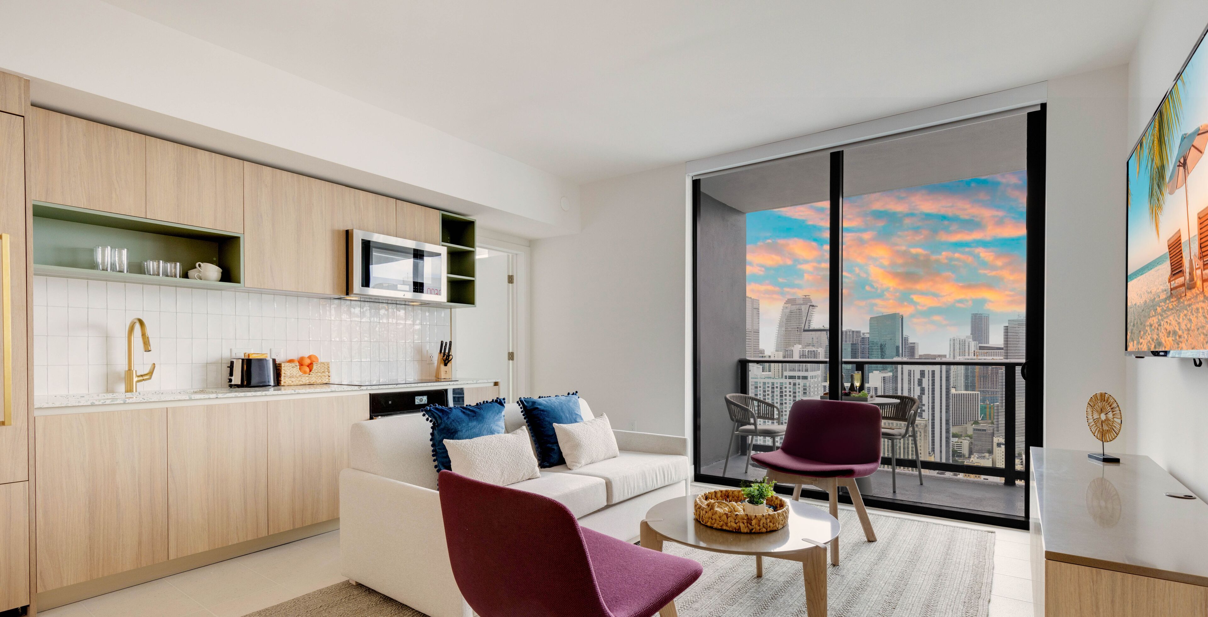 Una cocina moderna con armarios blancos, un microondas y una cafetera. Hay una zona de comedor con un sofá blanco y una silla morada. Un ventanal ofrece vistas a un horizonte urbano y al atardecer.