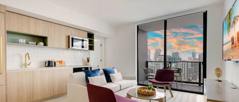 Una cocina moderna con armarios blancos, un microondas y una cafetera. Hay una zona de comedor con un sofá blanco y una silla morada. Un ventanal ofrece vistas a un horizonte urbano y al atardecer.