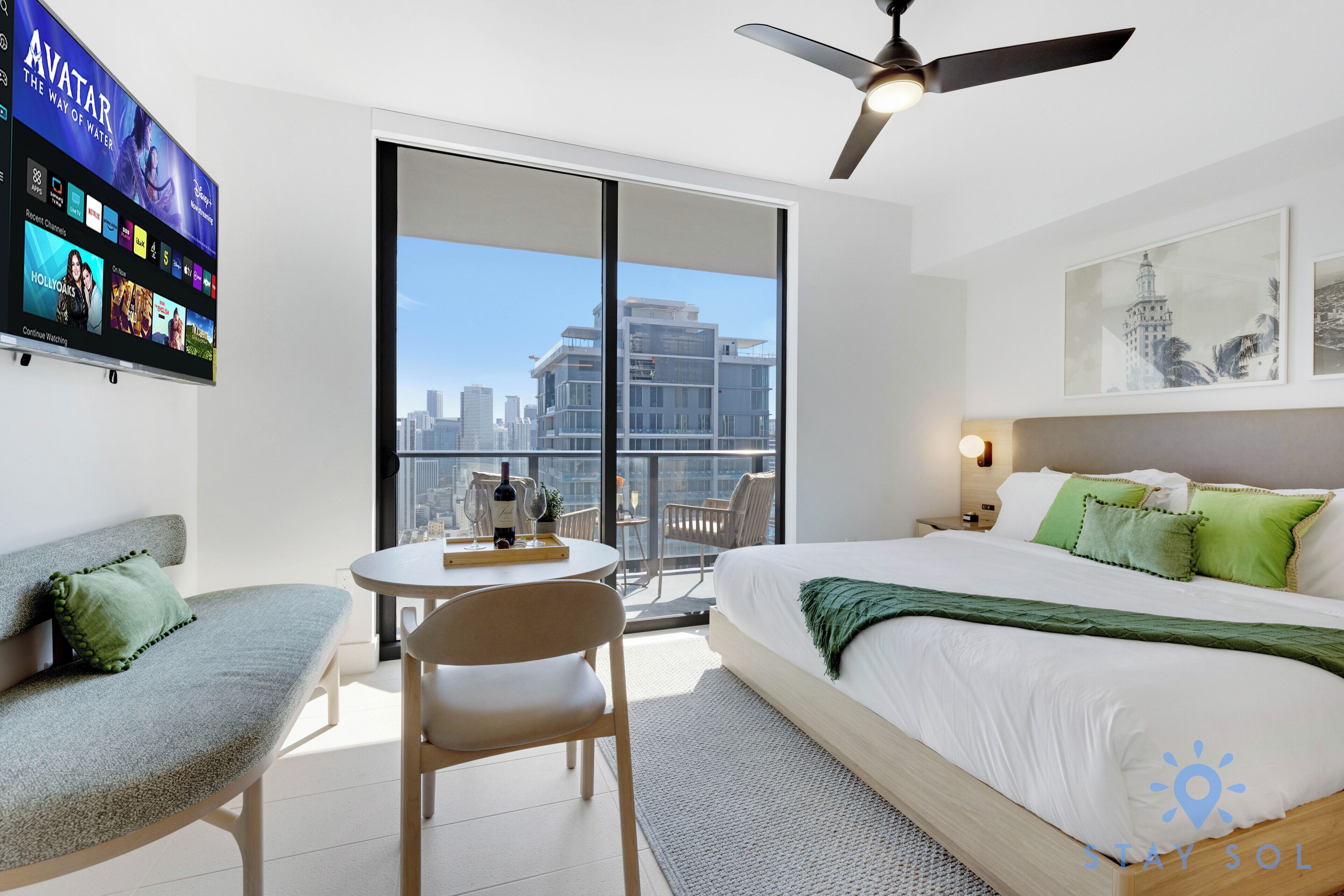 Una habitación de hotel moderna con una cama grande, un balcón con vista a la ciudad, una zona de comedor con mesa y sillas, y un televisor que muestra diferentes canales.