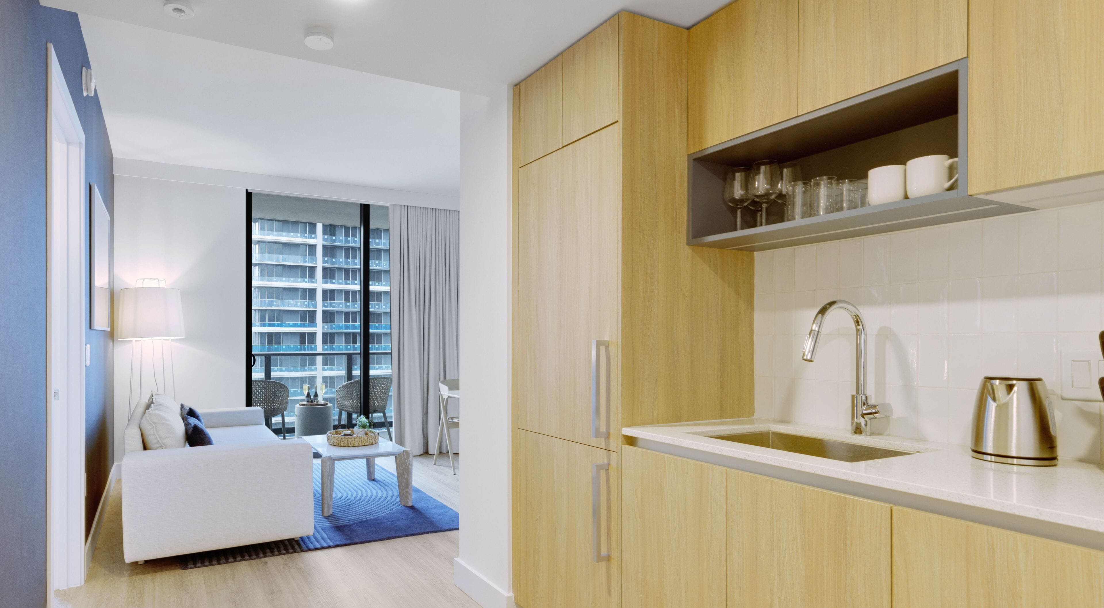 A modern hotel room with a kitchenette, a dining area with a view of a high-rise building, and a large window with curtains.