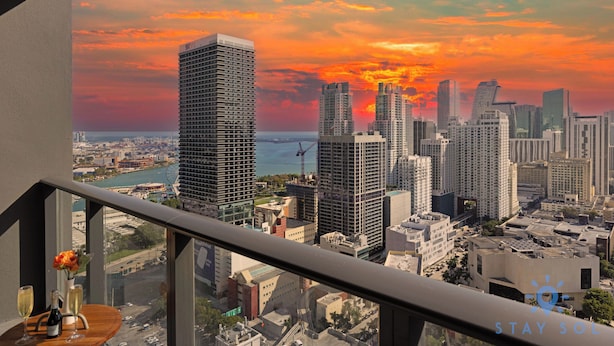 A cityscape view from a balcony with a sunset, featuring high-rise buildings and a waterfront. - Stay Sol Downtown (Miami)