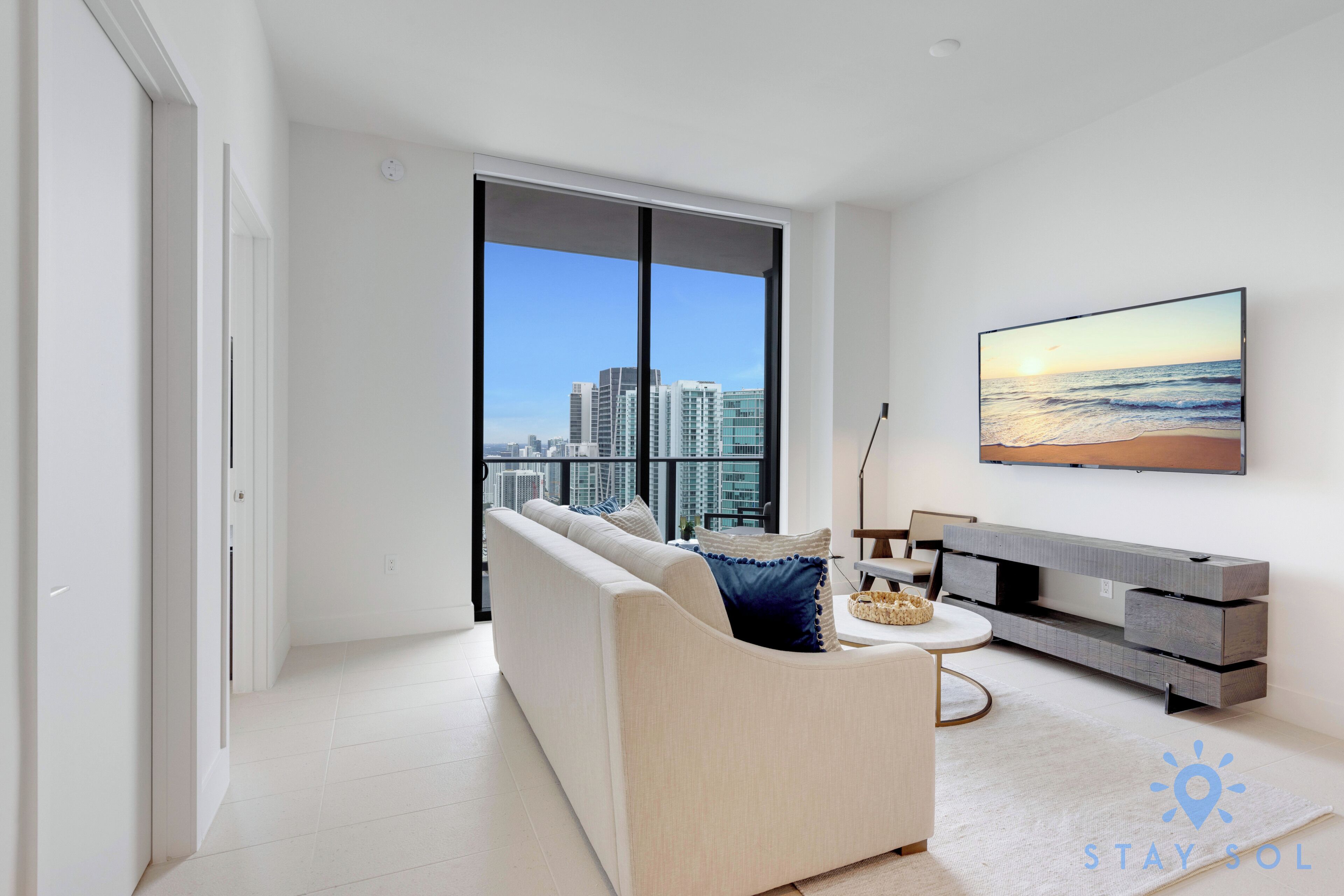 A modern living room with a large window, a sofa, a coffee table, and a flat-screen TV on the wall.