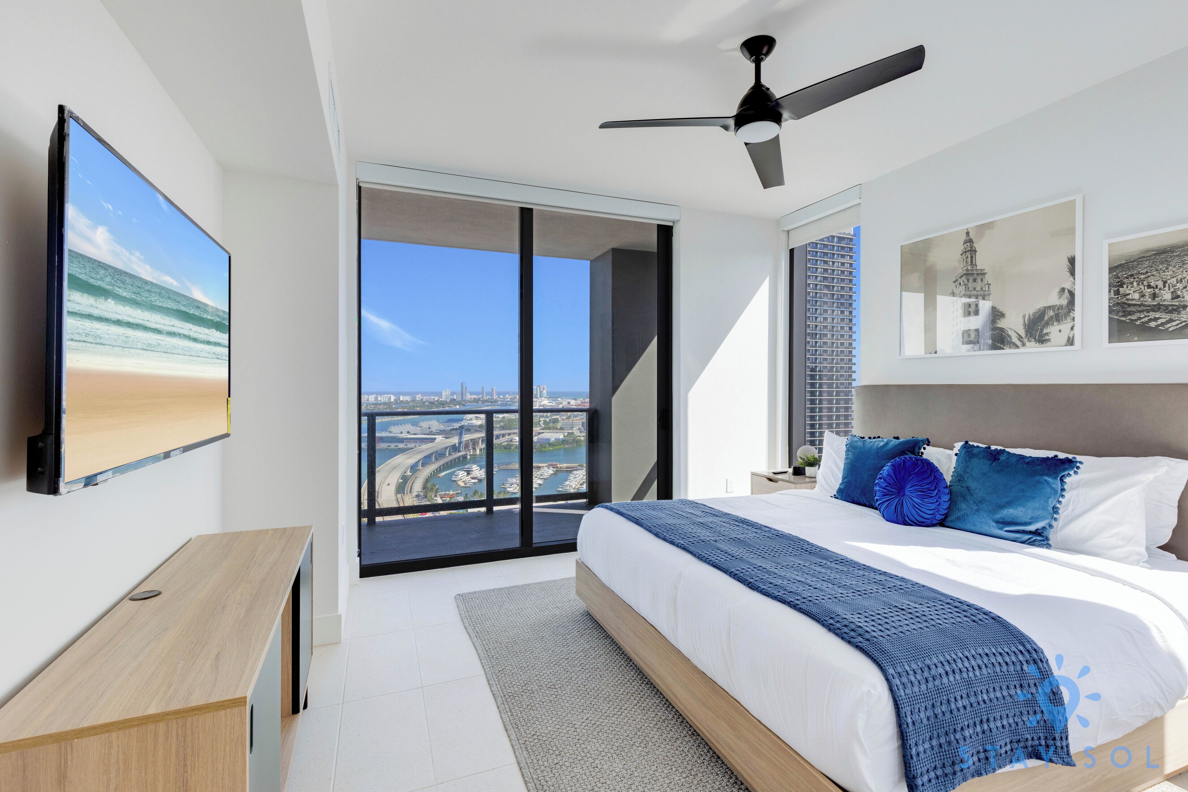 Une chambre moderne avec un grand lit, un ventilateur de plafond, une vue sur la ville et un téléviseur fixé au mur.