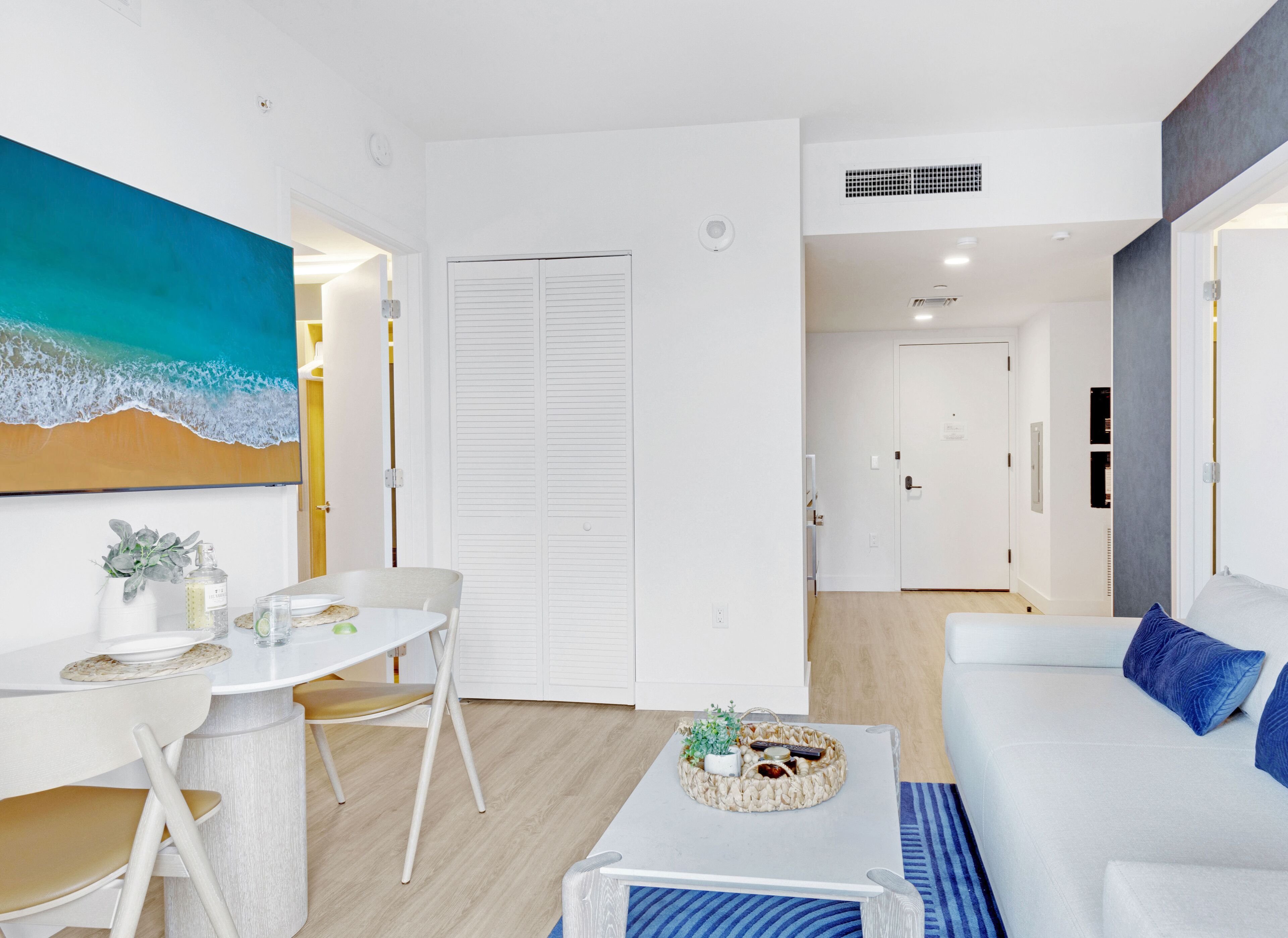Una sala de estar moderna con un sofá blanco, una mesa de comedor con sillas, una mesa baja con una cesta, y un cuadro de una escena de playa.