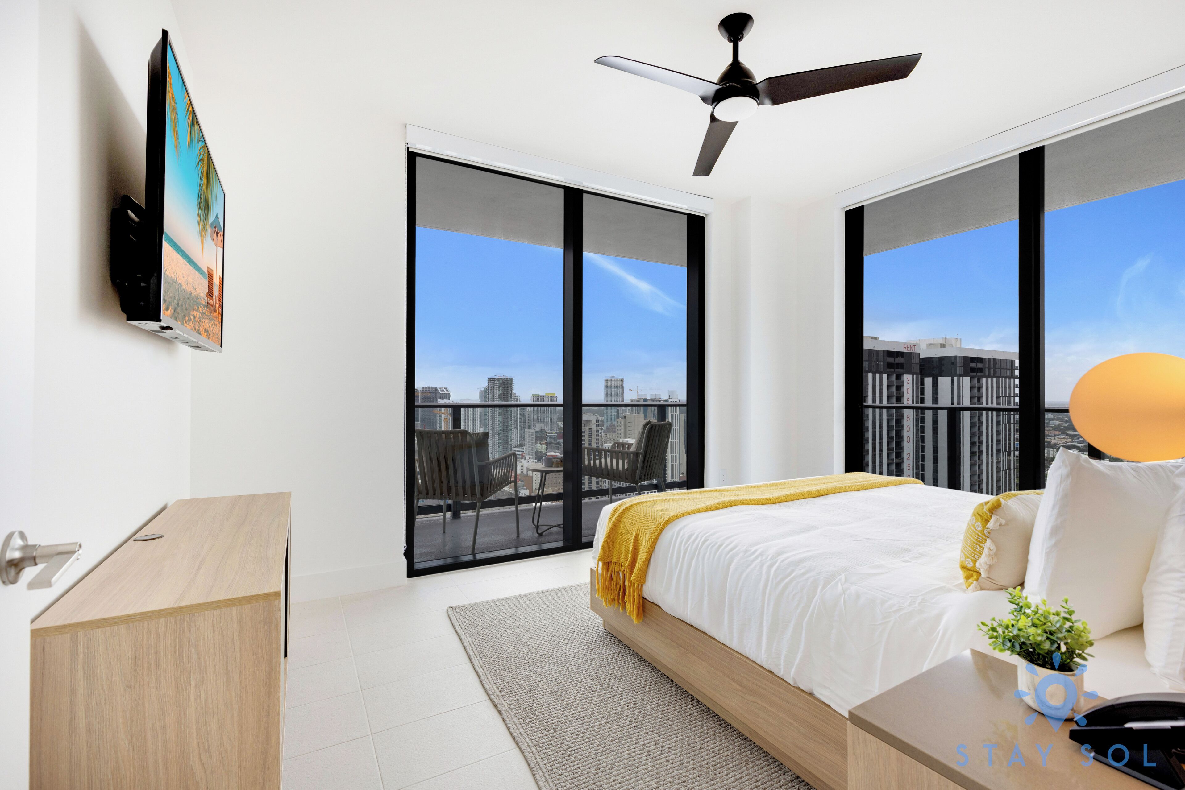 Ein modernes Hotelzimmer mit einem großen Bett, einem Schreibtisch, einem Deckenventilator und Blick auf die Stadt durch raumhohe Fenster.