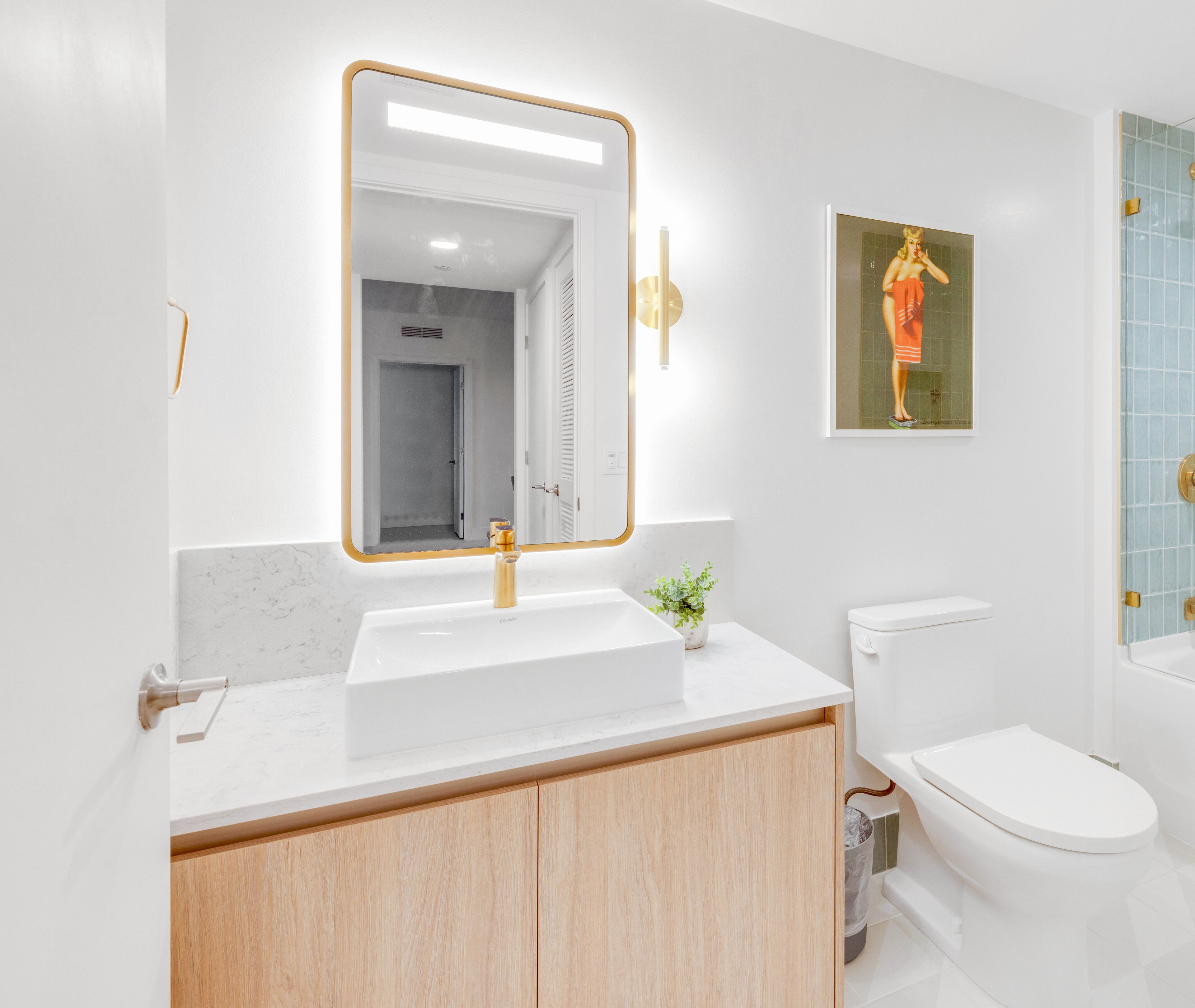 A modern bathroom with a white sink, a mirror with a gold frame, a toilet, and a small plant on the counter.