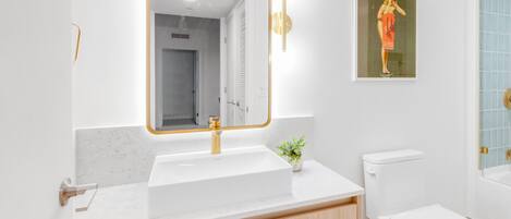 A modern bathroom with a white sink, a mirror with a gold frame, a toilet, and a small plant on the counter.