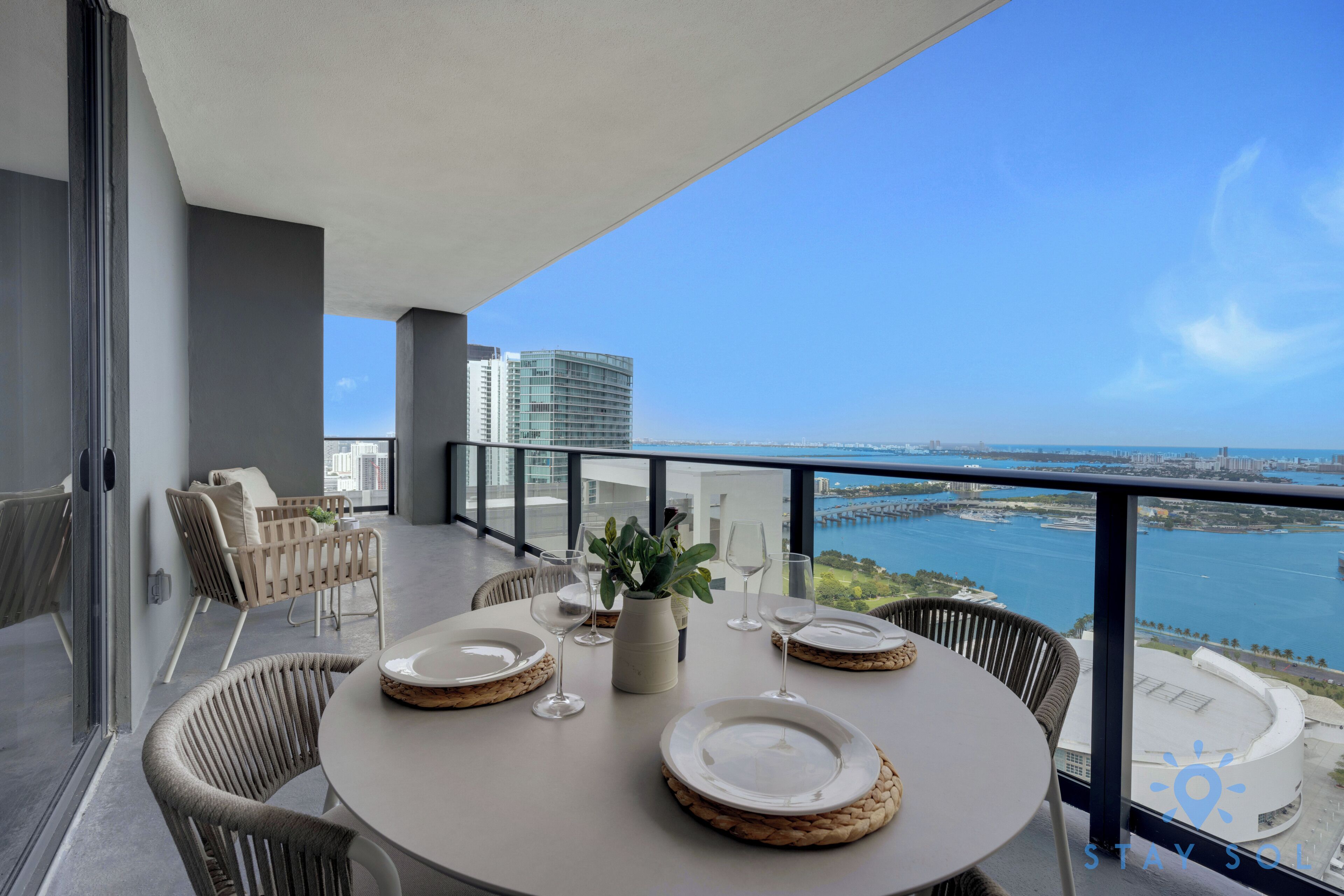 A balcony with a dining setup, including a round table, chairs, and a view of a body of water and cityscape.
