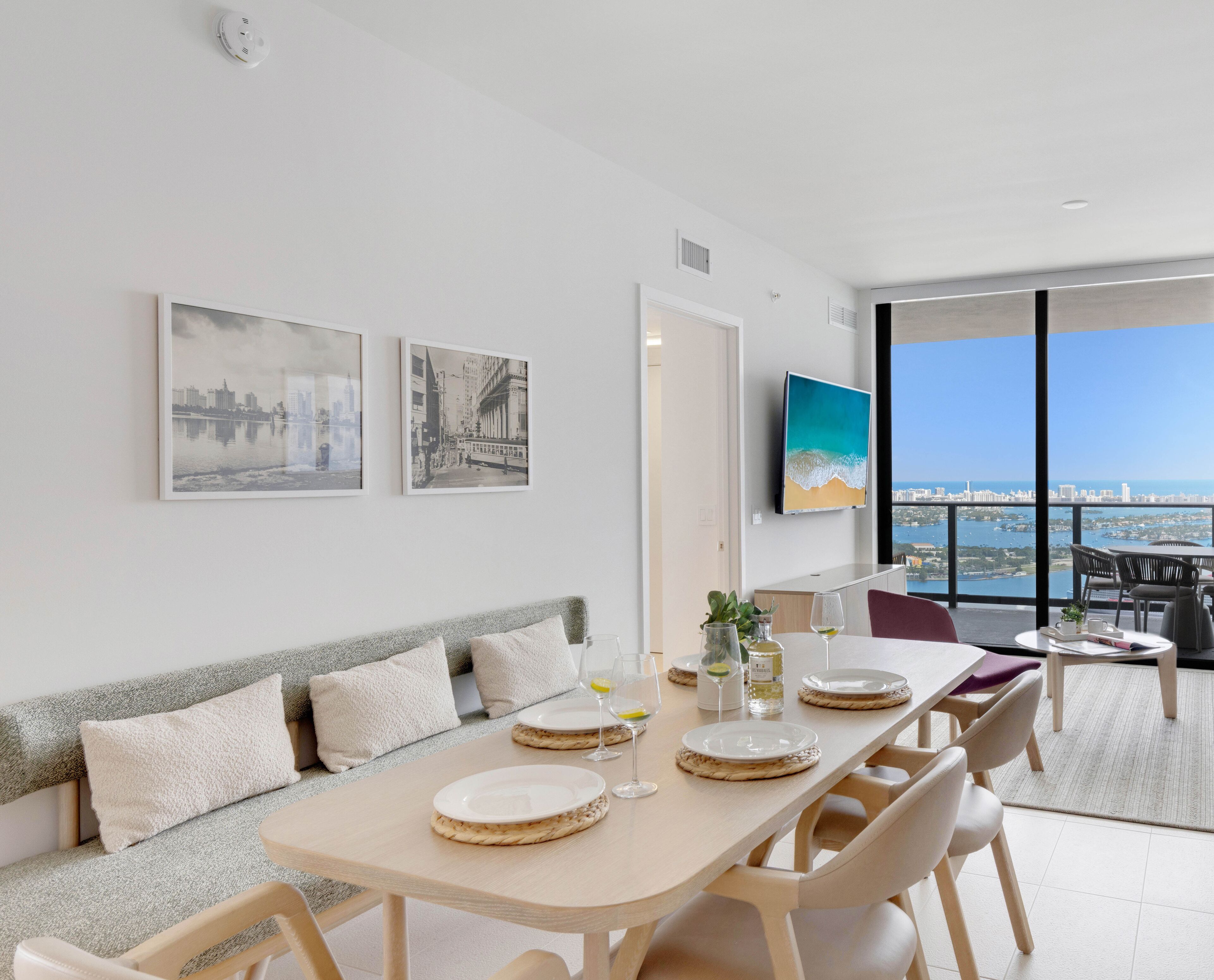 A modern dining area with a long table, chairs, and a view of the city and sea through large windows.