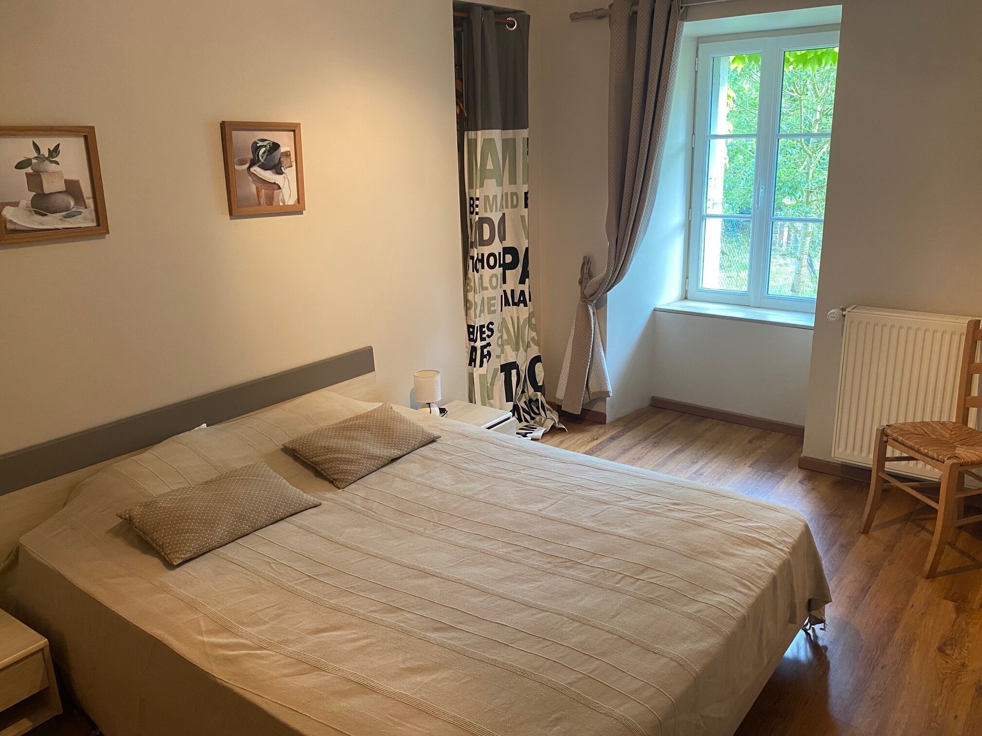 Une chambre avec un grand lit, une table de chevet, une chaise, une fenêtre avec des rideaux et deux tableaux encadrés accrochés au mur.