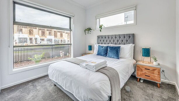 A bedroom with a large bed, a nightstand, a bedside table, a window with a view of construction, and a small mirror on the wall. - StayAU Fresh Modern 5BR Retreat Netflix in Berwick (Berwick)
