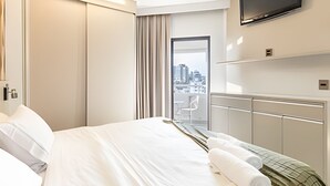 A hotel room with a bed, a TV mounted on the wall, a balcony with a view of the city, and a small table with towels.