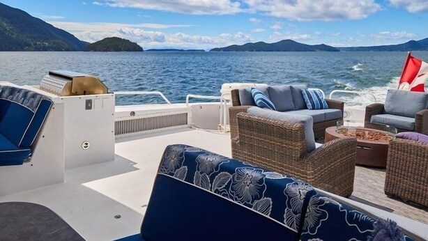 Cubierta de barco con mobiliario de mimbre, una bandera y vista al agua y las montañas.