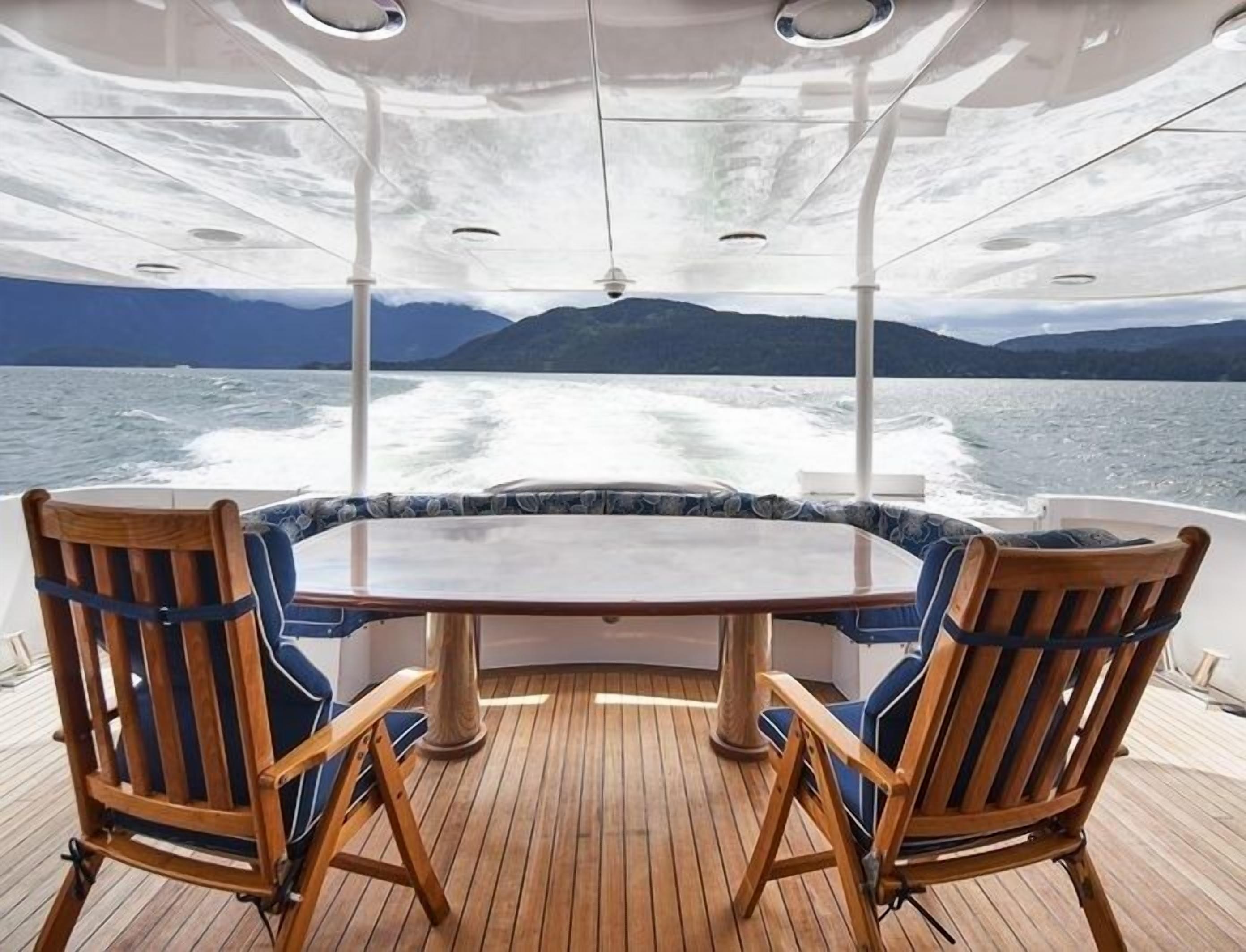 A boat deck with a round table and two wooden chairs, overlooking a body of water with waves and distant mountains.