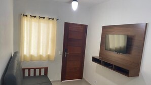 A room with a sofa, a wooden door, a TV mounted on the wall, and a ceiling fan. - Condomínio Gelatti (Ubatuba)