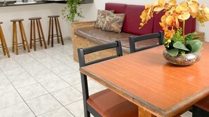 A dining area with a wooden table and chairs, a red sofa, and a potted plant with yellow flowers. - Condomínio Gelatti (Ubatuba)