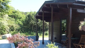 A covered outdoor patio with a brick planter, wooden chairs, and a hanging plant. - #2 Killarney Cabins, Stunning Lodge & Parking (Killarney)