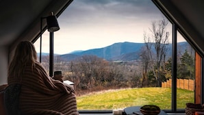 A cozy room with a large window overlooking a scenic landscape with mountains and trees. - Modern A-Frame w/ Hot Tub, Sauna & Stunning Views (Stanley)
