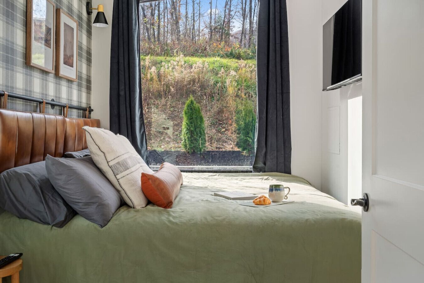 A bedroom with a bed, pillows, a bedside table, a window with curtains, and a view of trees and a hedge outside.