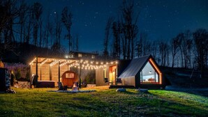 A modern house with a large circular wooden structure, a smaller cabin, and string lights. - Modern A-Frame w/ Hot Tub, Sauna & Stunning Views (Stanley)
