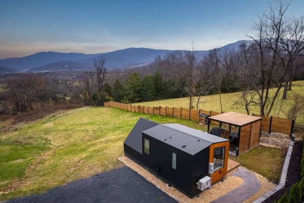 A modern cabin with a black exterior and a wooden deck, surrounded by a fenced yard and a view of distant mountains.
