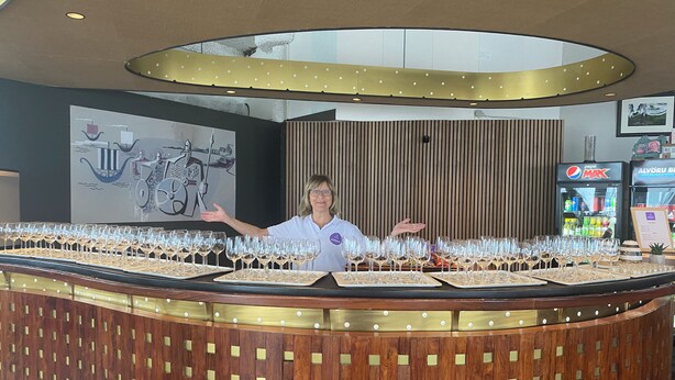 A bar area with multiple glasses of wine on trays, a Pepsi Max refrigerator, and a wooden wall with a vertical design. - Blue Hostel Bláu Fjöll (Vestmannaeyjar)