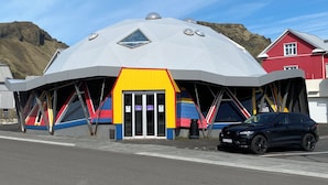 A geodesic dome structure with a colorful facade, a black car parked in front, and a red building in the background. - Blue Hostel Bláu Fjöll (Vestmannaeyjar)