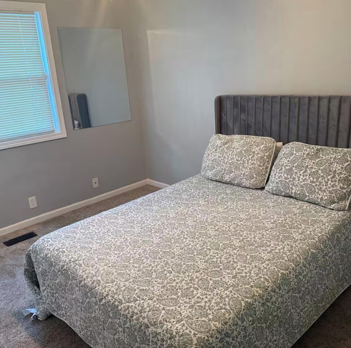 A bedroom with a bed, a window with blinds, a mirror, and a wall-mounted heater.