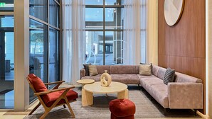 Um lobby moderno com uma grande janela de vidro, um sofá bege, uma mesa de centro de madeira e um banquinho vermelho.