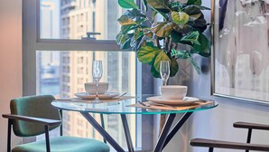 Uma área de refeições com mesa de vidro, duas cadeiras, um vaso com planta e vista para a cidade pela janela.