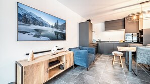 A modern living room with a large TV, a blue sofa, a wooden cabinet, and a kitchen area in the background. - Rivulet - Comfortable apartment in Châtel (Châtel)