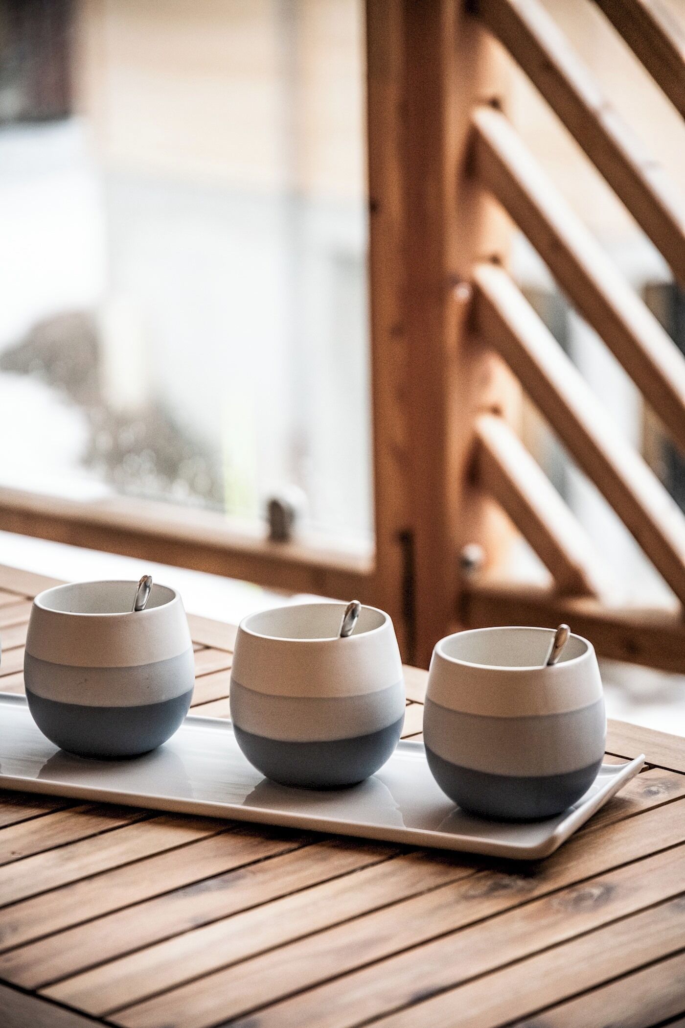 Three ceramic cups on a wooden tray on a table by a window with a wooden railing.