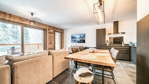 A modern interior with a wooden dining table, cushioned seating, a kitchen area, and large windows offering a view of trees. - Rivulet - Comfortable apartment in Châtel (Châtel)