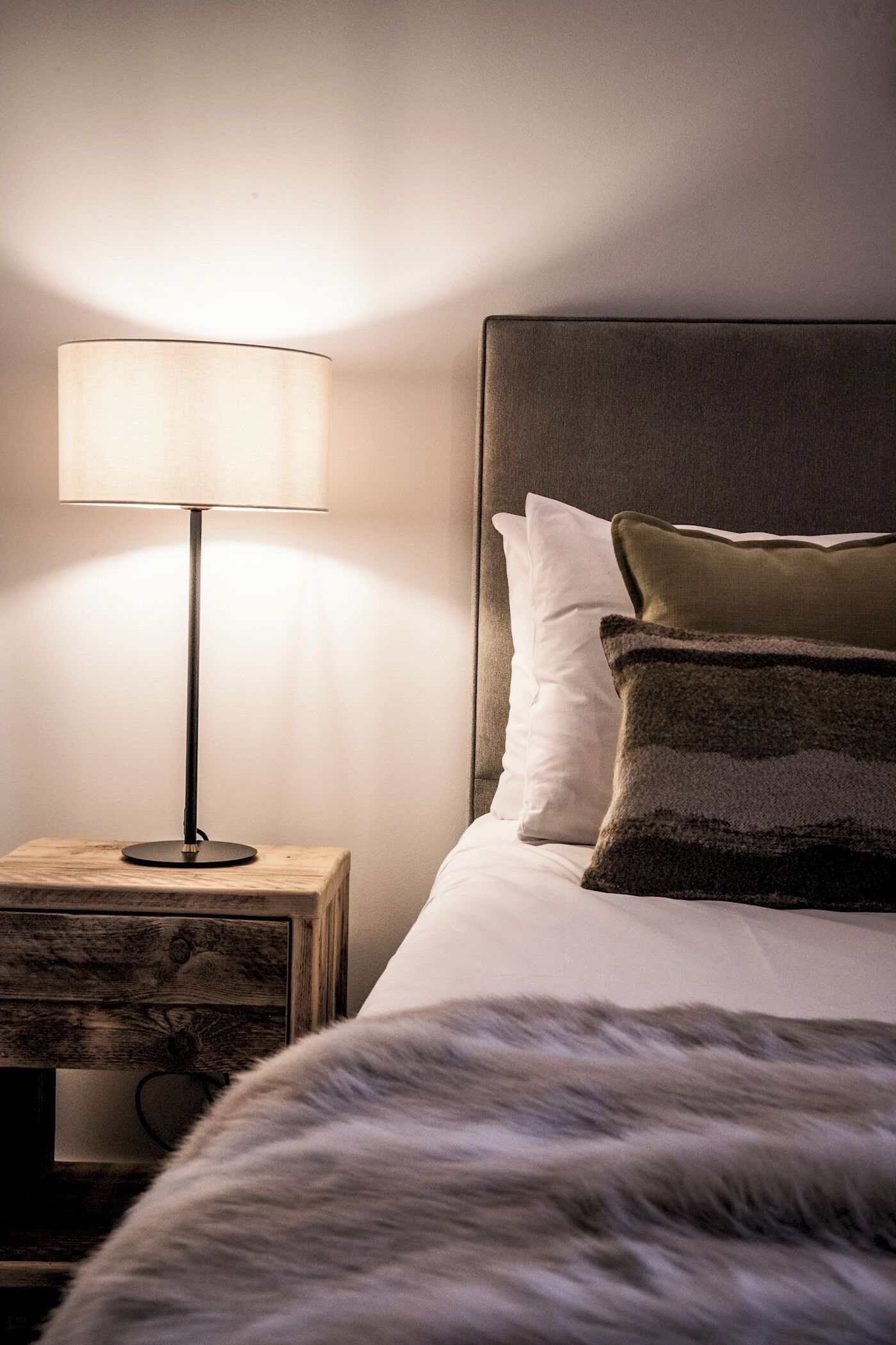A neatly made bed with white and dark pillows, a wooden nightstand with a lamp, and a headboard against a plain wall.