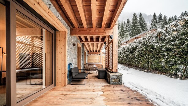 A wooden deck with a glass door leading to a room, a seating area with chairs, and a snowy landscape with trees in the background. - Ubuntu - Stylish Alpine Apartment in Châtel (Châtel)