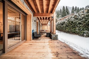 Een houten terras met een glazen deur die naar een kamer leidt, een zithoek met stoelen, en een besneeerd landschap met bomen op de achtergrond.