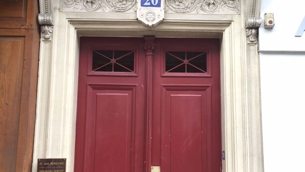Une porte rouge à deux battants, surmontée d’une plaque portant le numéro « 20 », et encadrée de moulures blanches décoratives.